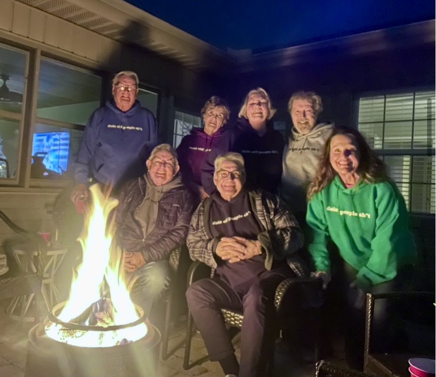 Seven friends gathered around a backyard fire pit at night in DOPS.LIFE “doin old people sh*t” hoodies — navy, burgundy, black, gray and green