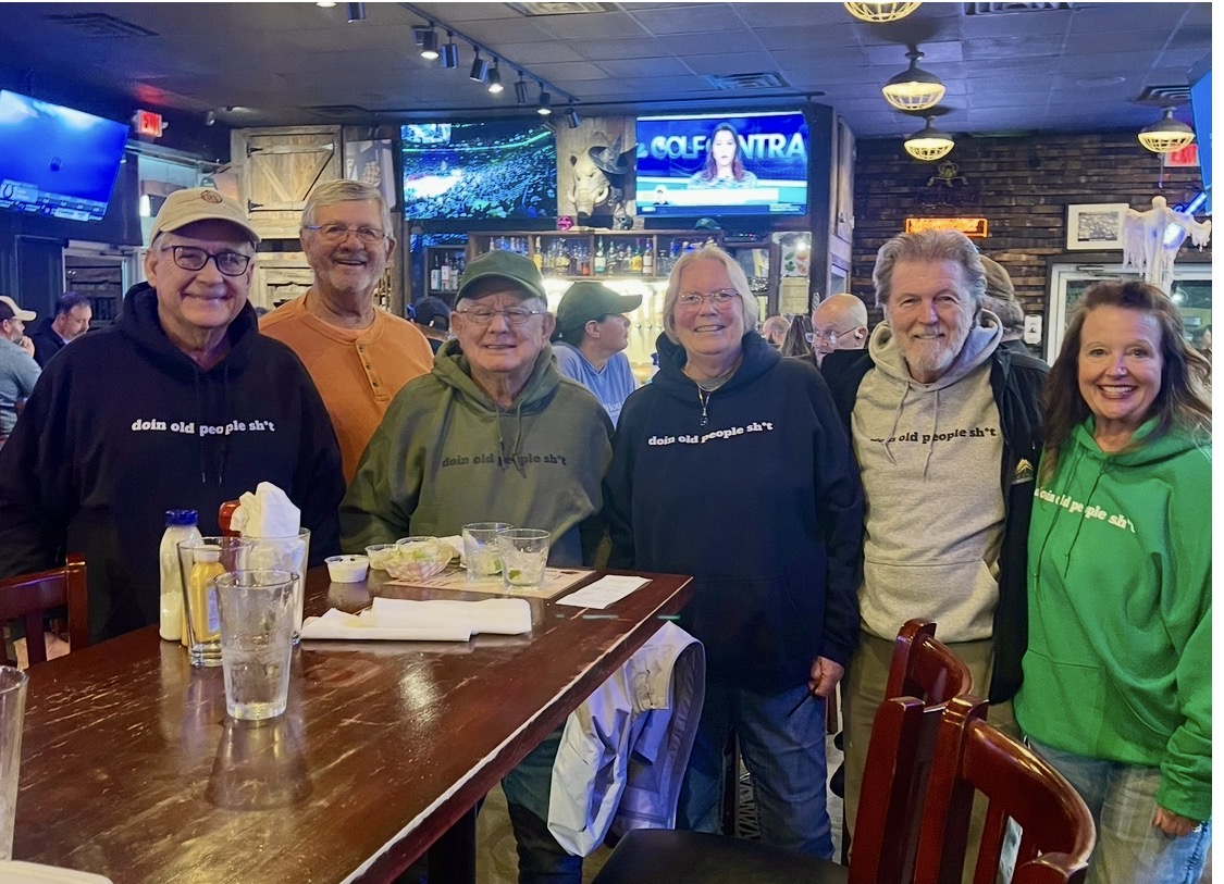 Friends at a tavern wearing DOPS.LIFE “doin old people sh*t” hoodies in black, green, orange and gray